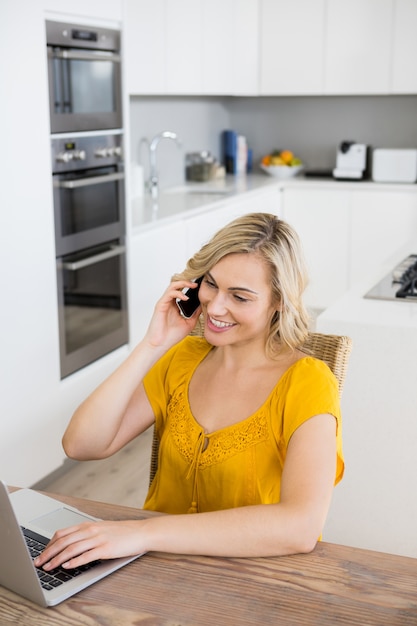 Женщина разговаривает по мобильному телефону во время использования ноутбука на кухне