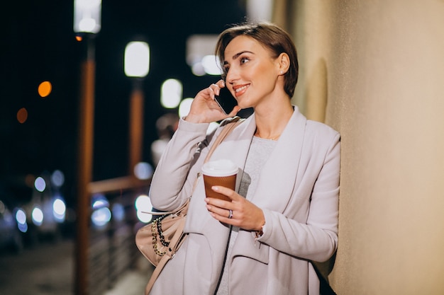Woman talking on phone and drinking coffee outside in the street at night