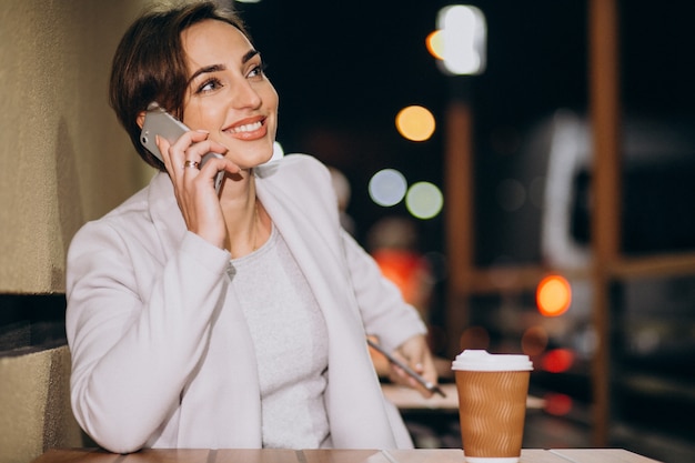 女性が電話で話していると夜に路上で外のコーヒーを飲む