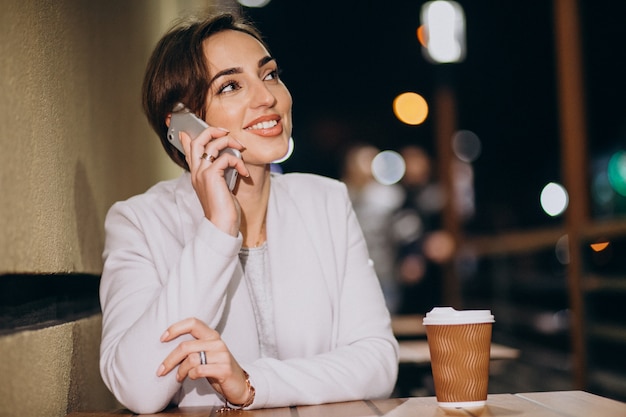 女性が電話で話していると夜に路上で外のコーヒーを飲む