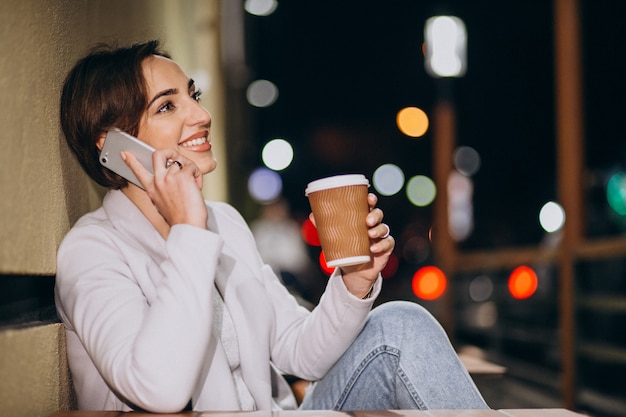 女性が電話で話していると夜に路上で外のコーヒーを飲む