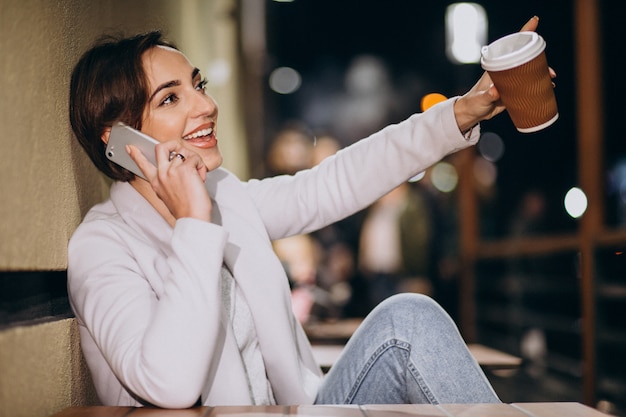 Woman talking on phone and drinking coffee outside in the street at night