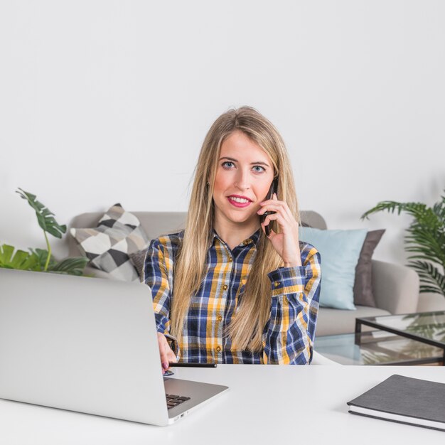 Woman talking on phone while sitting at table with laptop 