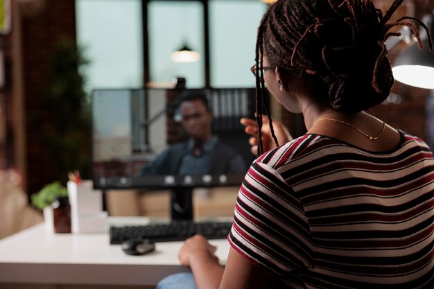 Женщина разговаривает с сотрудником по видеоконференции, афро-американский фрилансер, работающий дома. Удаленный работник посещает виртуальную встречу, общается на телеконференции с помощью компьютера