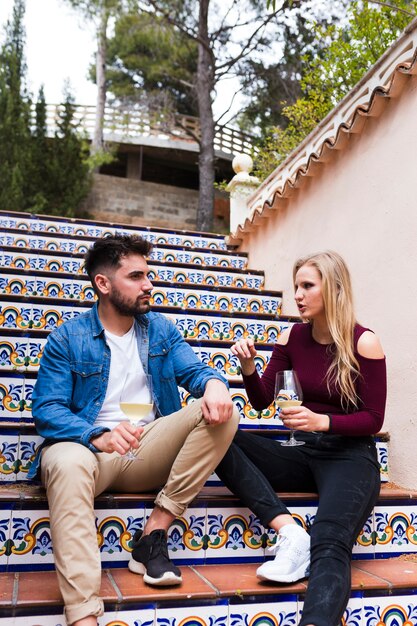 Woman talking with her male friend while sitting on staircase with glass of alcohol