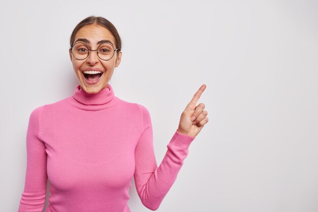 woman talks about personal achievements points aside at upper right corner checks out awesome discount promotion wears pink poloneck and spectacles poses indoor