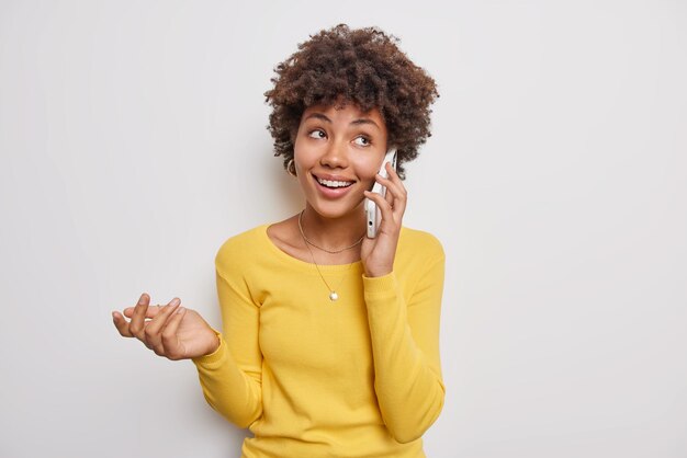 woman talks via smartphone with friend gestures with hand and smiles positively makes phone call wears casual yellow sweater isolated on white. Communication.