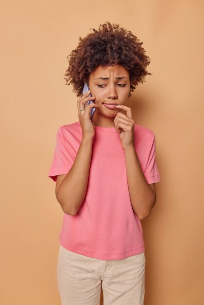 woman tas telephone conversation hears bad news looks down with gloomy worried expression wears casual pink t shirt and trousers isolated on beige