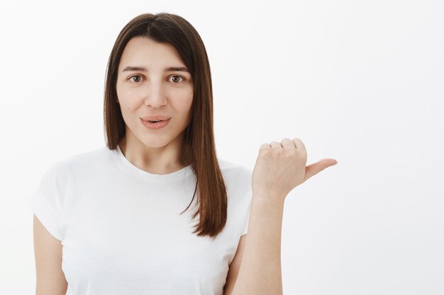 Woman telling about awesome product indicating with thumb right to take look yourself smiling, folding lips from amazement and interest recommending watch and try, posing against white wall