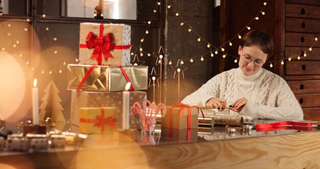 Woman in thick white cable sweater wrapping presents on the background of Christmas decorations and lights