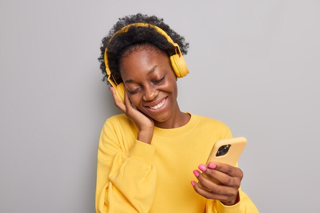  woman tilts smiles pleased listens music in wireless headphones holds smartphone dressed in yellow jumper isolated on grey 