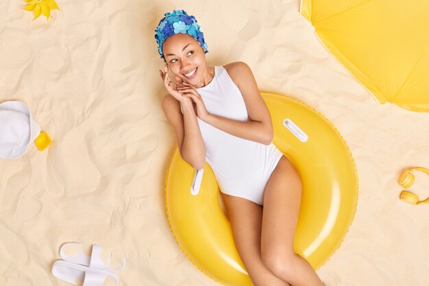 woman touches face gently looks away happily wears blue headwear white swimsuit sits on yellow inflated swimring has dreamy expression relaxes at beach