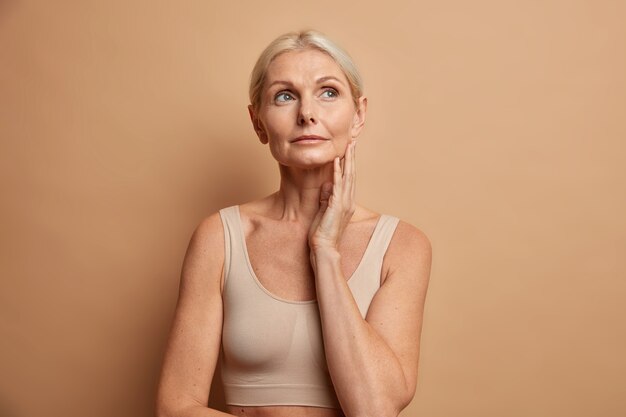 woman touches skin after applying anti age cream concentrated above with thoughtful expression wears cropped top isolated on brown