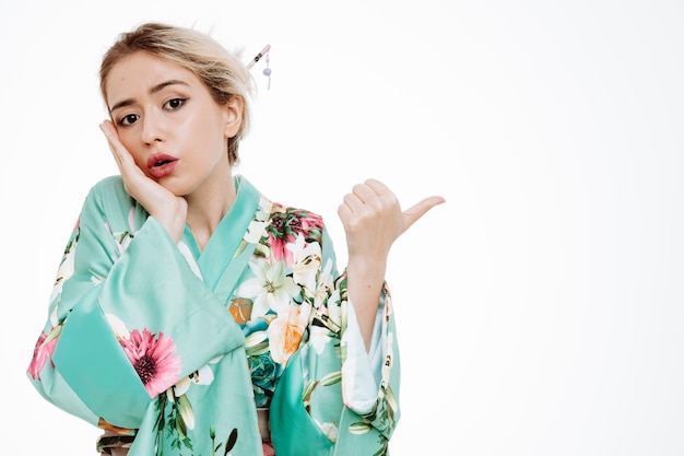 Woman in traditional japanese kimono being surprised pointing with thumb to the side on white