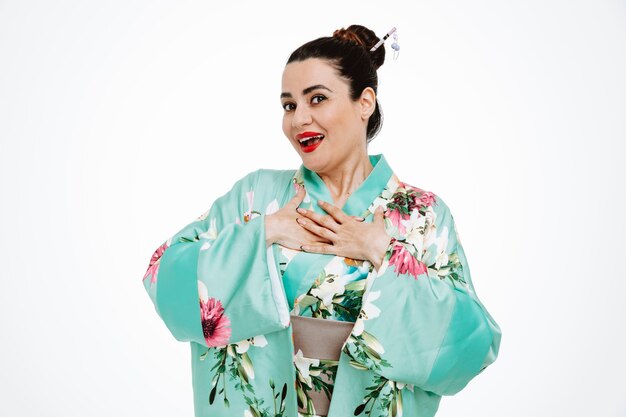 Woman in traditional japanese kimono happy and cheerful holding hands on herchest feeling thankful on white