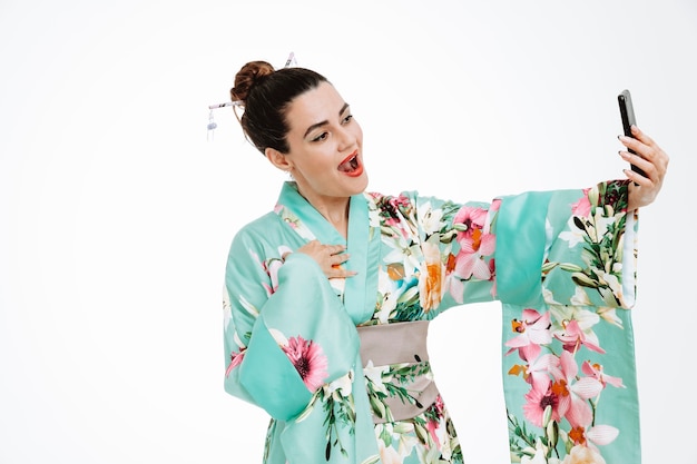 Woman in traditional japanese kimono happy and positive smiling doing selfie using smartphone on white