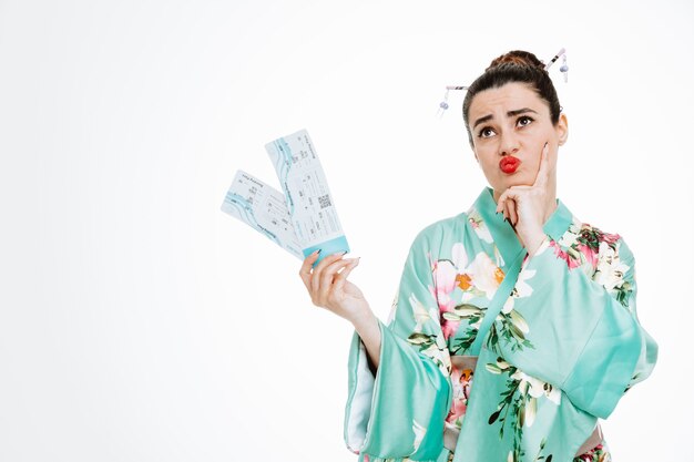 Женщина в традиционном японском кимоно, держащая авиабилеты, смотрит вверх с задумчивым выражением лица, держась за подбородок на белом