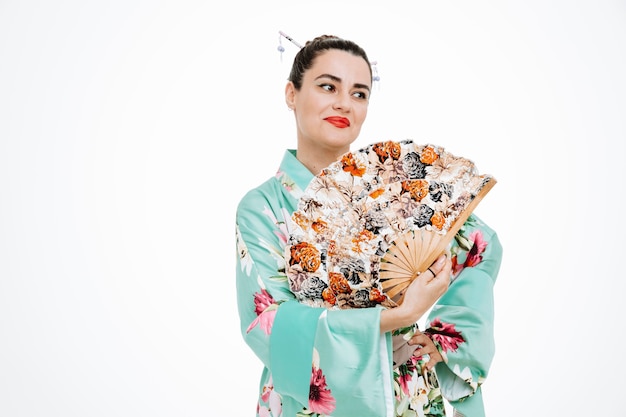 Woman in traditional japanese kimono holding hand fan looking aside happy and cheerful smiling on white