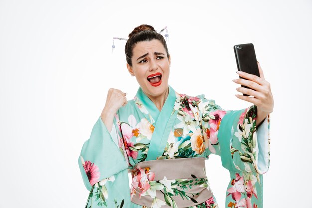 Woman in traditional japanese kimono holding smartphone clenching fist rejoicing her success crazy happy and excited on white