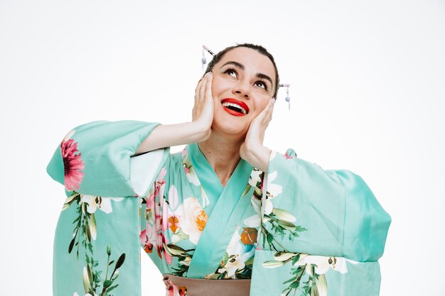 Женщина в традиционном японском кимоно смотрит вверх счастливой и удивленной, мечтая держась за щеки, широко улыбаясь на белом