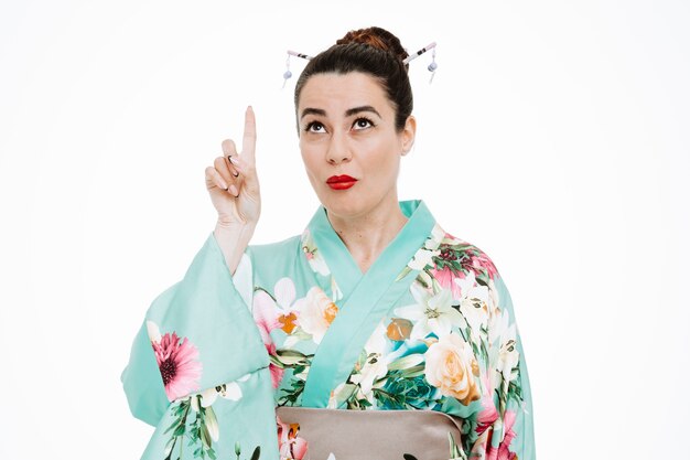 Woman in traditional japanese kimono looking up pointing with index finger up having doubts being confused on white