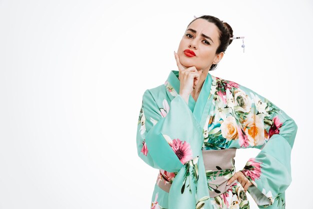 Woman in traditional japanese kimono with skeptic expression holding hand on her chin on white