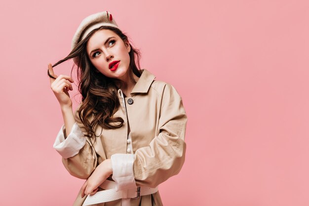 Woman in trench coat and beret looks thoughtfully up, bites her lip and winds her hair onto her finger.