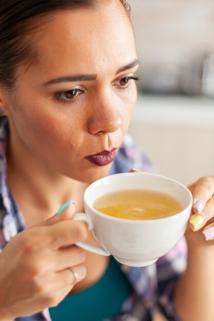 Woman trying to drink hot green tea with aromatic herbs in kitchen in the morning