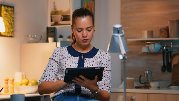 Woman typing on tablet working from home sitting on chair in the kitchen late at night. Busy focused employee using modern technology network wireless doing overtime writing, searching.