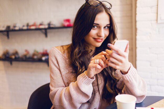 Женщина, набрав написать сообщение на смарт-телефон в современном кафе. Подрезанное изображение молодой милой девушки сидя на таблице с кофе или капучино используя мобильный телефон.