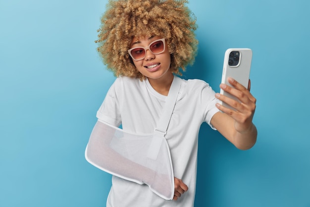 Woman uses cellular for taking selfie wears casual white t shirt splint on broken arm wears sunglasses and white t shirt isolated over blue background makes video call tells friend about accident