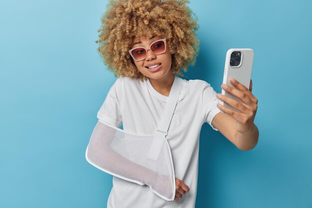 Женщина использует мобильный телефон для селфи, носит повседневную белую футболку с шиной на сломанной руке, носит солнцезащитные очки и белую футболку, изолированную на синем фоне, делает видеозвонок и рассказывает другу об аварии