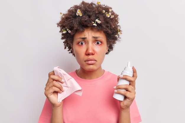 여성은 비강 에어로졸을 사용하여 알레르기성 비염으로 고통받고 있습니다. 빨간색으로 부어오른 눈은 흰색 위에 격리된 캐주얼 티셔츠를 입은 불행해 보입니다.