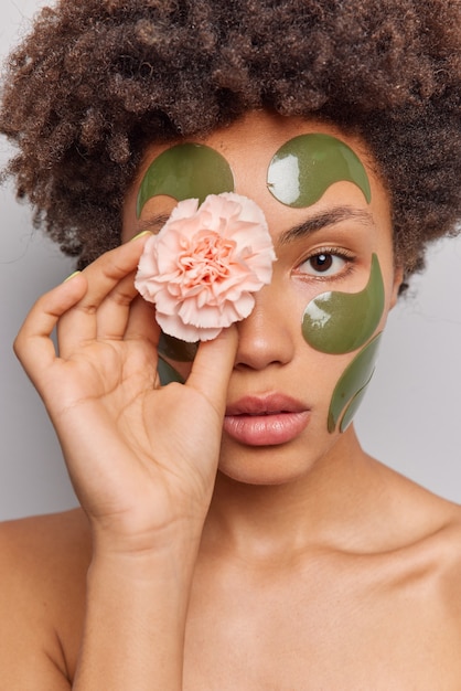 женщина использует натуральные косметические средства, держит цветок на глазах, применяет коллагеновые зеленые пятна на лице, стоит без рубашки, позы в помещении