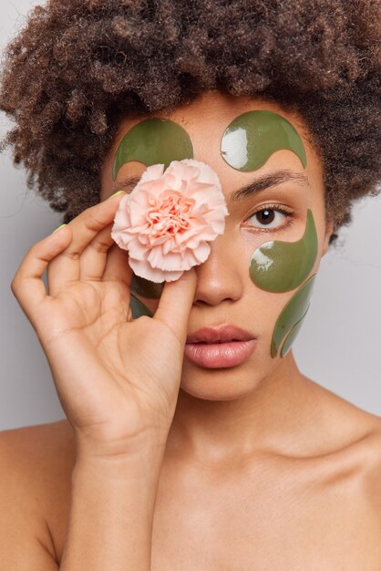 woman uses natural beauty products holds flower on eye applies collagen green patches on face stands shirtless poses indoor