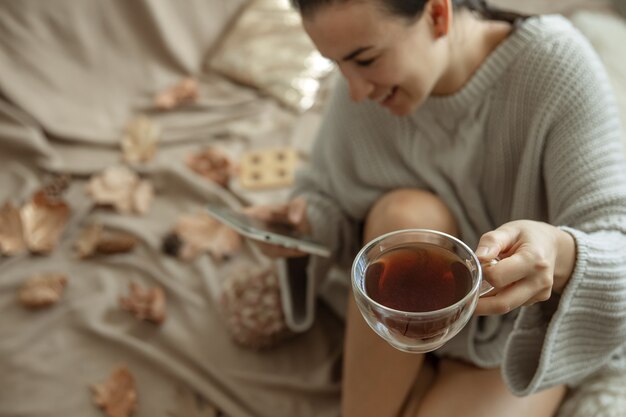a woman uses the phone and holds a cup of tea while sitting in bed in a cozy knitted sweater, blurred background.
