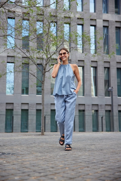  woman uses roaming connection for communication walks around city calls operator for checking account balance wears blue summer costume and sandals