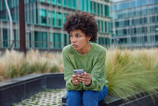 woman uses smartphone browses web pages chats with followers at own wrbsite uses application on mobile phone communicates and reads news poses outdoors against urban buildings