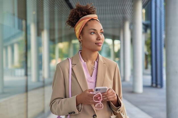 woman uses smartphone while strolls in urban setting holds sunglasses dressed in stylish clothes looks away sends text messages to boyfriend