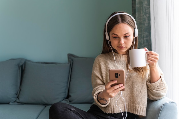 Woman using headphones and smartphone at home while having coffee during the pandemic