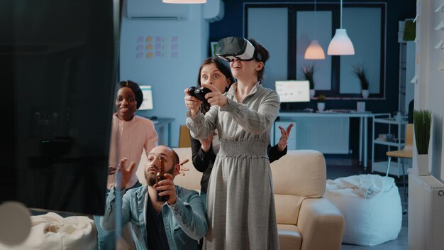 Woman using joystick and vr glasses while workmates cheering after work. Coworkers enjoying video games to play game on tv console with virtual reality goggles and controller at office