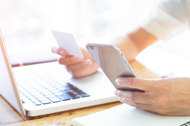 Женщина, используя ноутбук и мобильный телефон для онлайн-покупок и оплаты кредитной картой. Эта фотография фокусируется на руке женщины и использует теплый яркий солнечный фильтр для комфортного ощущения