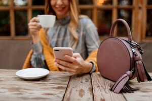 Free photo woman using mobile phone, texting message and drinking coffee. stylish bag on table. wearing grey dress and orange plaid. enjoying cozy morning in cafe.