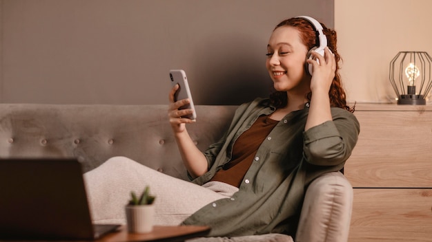 Foto gratuita donna che utilizza cuffie moderne e smartphone sul divano di casa