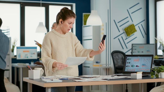 Woman using smartphone in video call conference talking about sales holding paper with charts in busy startup office. Business employee talking with remote coworker using mobile phone.