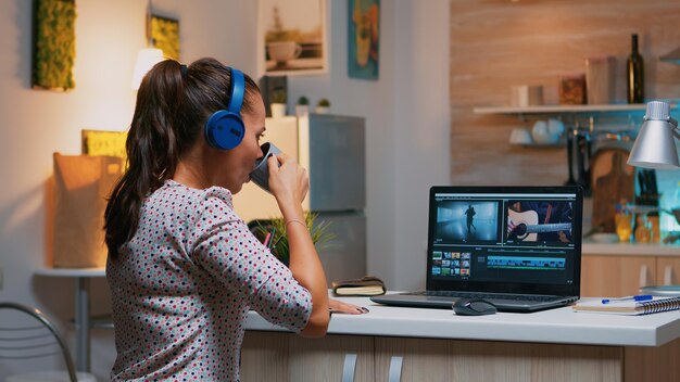 Woman video editor with headset working with footage and sound sitting in home kitchen. Woman videographer editing audio film montage on professional laptop sitting on desk in midnight