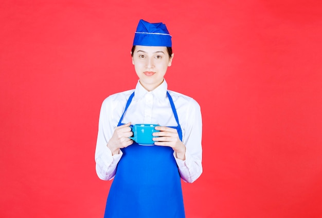 Cameriera donna in uniforme in piedi e con in mano una tazza blu sul muro rosso.