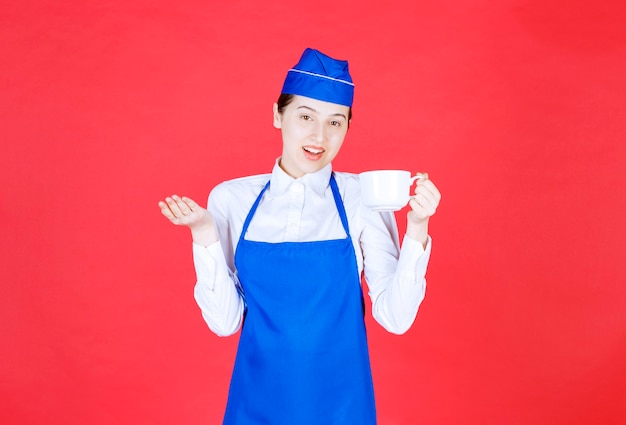 Cameriera donna in uniforme in piedi e con in mano una tazza sul muro rosso.