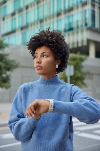 woman waits for someone at street checks time onn wristwatch wears casual blue jumper strolls in downtown poses outside considers something