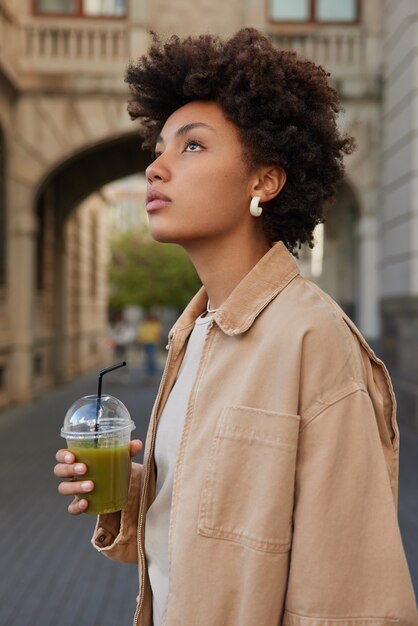woman walks in city alone drinks fresh smoothie looks above strolls at urban place dressed in beige jacket